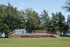 An outdoor stage constructed with logs and rustic wooden boards at the edge of a lake.